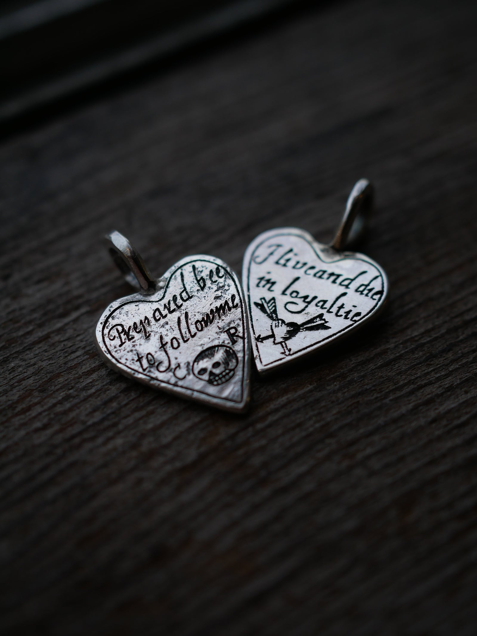 Two silver heart-shaped pendant charms with engraved text and symbols, displayed on a dark wooden surface.