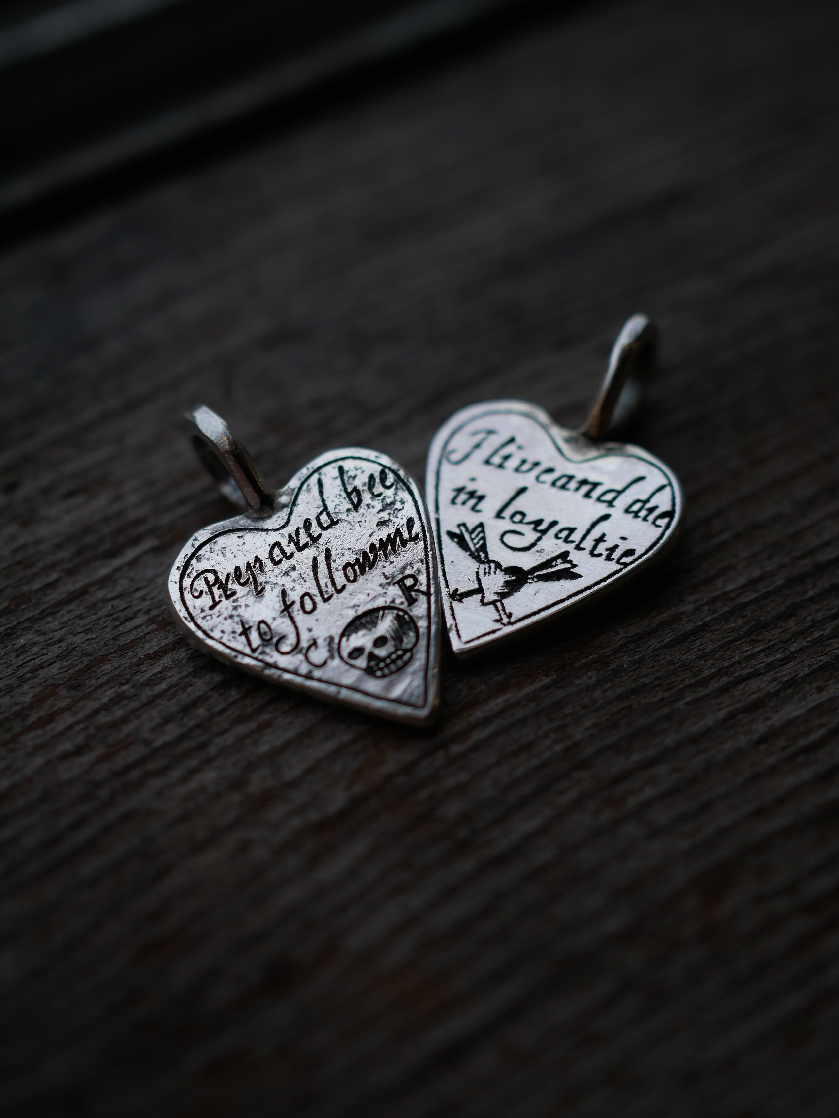 Two silver heart-shaped pendant charms with engraved text and symbols, displayed on a dark wooden surface.